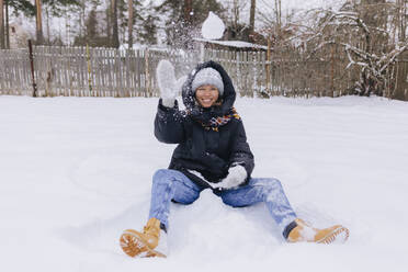 Happy woman sitting on snow field playing with snowballs - KNTF04193