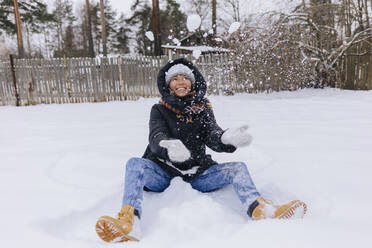 Happy woman sitting on snow field playing with snowballs - KNTF04194