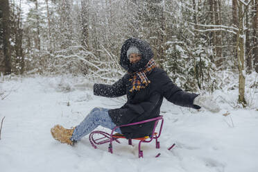 Smiling woman sitting on sledge throwing snow into the air in winter forest - KNTF04230