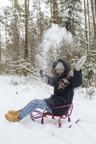 Glückliche Frau sitzt auf einem Schlitten und wirft Schnee in die Luft im Winterwald, lizenzfreies Stockfoto
