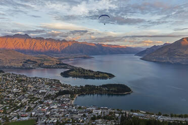 New Zealand, Otago, Queenstown, Paraglider flying over lakeshore town at dusk - FOF11808