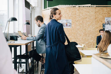 Smiling businesswoman with bag looking away while standing amidst colleagues in coworking space - MASF16304