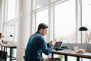 Junger Geschäftsmann, der ein Smartphone benutzt, während er mit einem Laptop am Schreibtisch im Büro sitzt - MASF16309