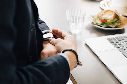 Mittelteil eines Geschäftsmannes, der seine Smartwatch überprüft, während er mit seinem Laptop am Schreibtisch im Büro sitzt - MASF16320