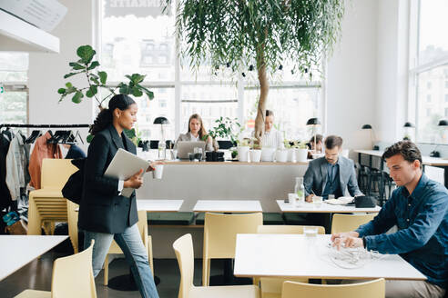 Geschäftsfrau mit Laptop, die spazieren geht, während Kollegen am Schreibtisch im Coworking Space arbeiten - MASF16325