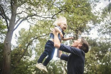 Low angle view of smiling man catching blond baby boy against trees - MASF16339