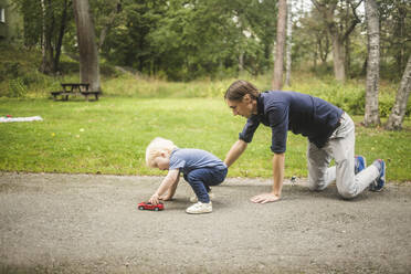 Vater kniet, während sein Sohn mit einem Spielzeugauto auf dem Gehweg vor Pflanzen im Park spielt - MASF16344