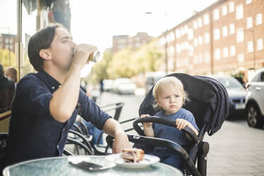 Ein kleiner Junge schaut weg, während sein Vater im Kinderwagen sitzt und in einem Straßencafé in der Stadt trinkt - MASF16359