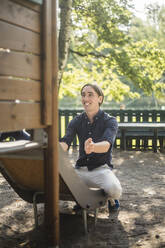 Lächelnder Mann, der auf einen kleinen Jungen wartet, während er auf einer Rutsche auf einem Spielplatz im Park sitzt - MASF16362