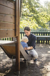 Smiling man holding baby boy while sitting on slide at playground in park - MASF16363