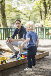 Man sitting while looking at son standing in playground - MASF16369