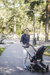 Ganzkörperporträt eines lächelnden Mannes, der mit seinem Sohn im Kinderwagen in einem öffentlichen Park steht - MASF16371