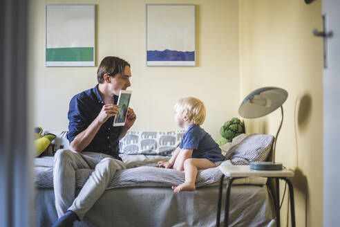 Mann zeigt seinem Sohn, der in der Wohnung im Schlafzimmer sitzt, ein Foto auf einem digitalen Tablet - MASF16376