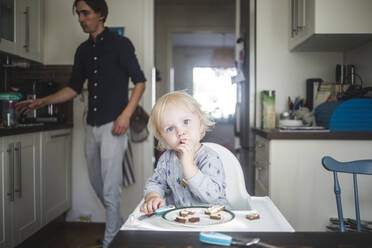 Porträt eines blonden kleinen Jungen, der auf einem Hochstuhl in der Küche sitzt, mit seinem Vater im Hintergrund - MASF16386
