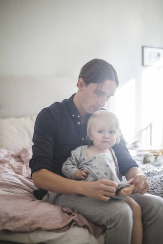 Vater, der einem kleinen Jungen beim Tragen von Kleidung hilft, während er zu Hause auf dem Bett sitzt, lizenzfreies Stockfoto