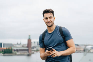 Porträt eines jungen Mannes, der sein Handy in der Hand hält, während er vor einer Stadtlandschaft am Himmel steht - MASF16395