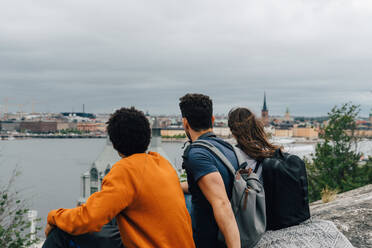 Rear view of friends sitting on hill by river in city against sky - MASF16398