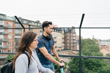 Männliche und weibliche Freunde benutzen Roller auf einer Brücke in der Stadt gegen den Himmel - MASF16407