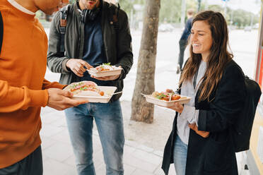 Smiling young woman enjoying meal with male friends while standing on street by food truck - MASF16415