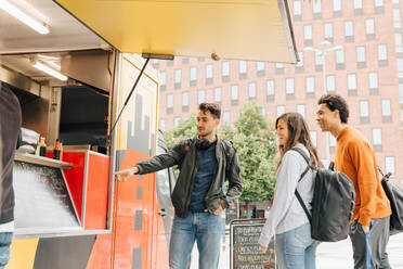 Young man pointing at menu while standing with friends by food truck in city - MASF16422