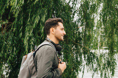 Seitenansicht eines jungen Mannes mit Rucksack, der an einem Baum in der Stadt steht, lizenzfreies Stockfoto