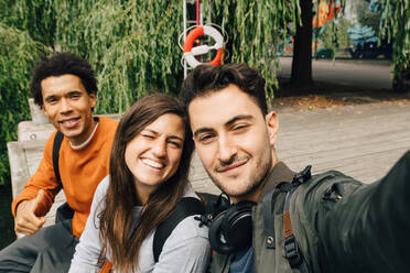 Portrait of smiling friends taking selfie while sitting on pier in city - MASF16427