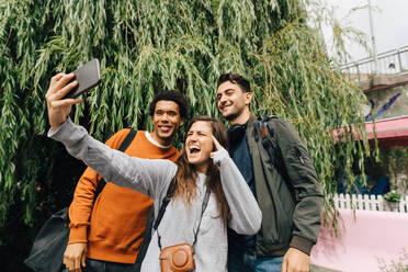 Lächelnde Freunde, die ein Selfie mit ihrem Mobiltelefon machen, während sie an einem Baum in der Stadt stehen - MASF16428