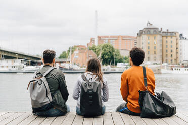 Rear view of friends with backpacks sitting on pier by river in city against sky - MASF16429