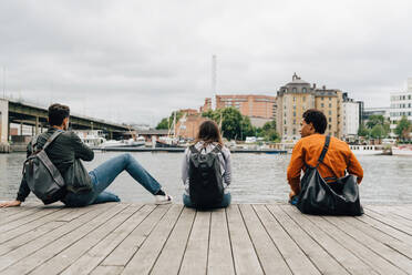 Rear view of friends sitting on pier by river in city against sky - MASF16430