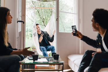 Man photographing friend with smart phone while sitting in swing at home - MASF16439