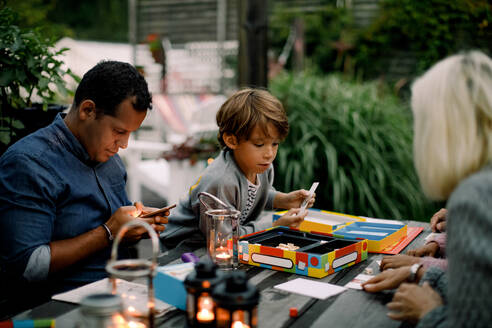 Familie spielt ein Brettspiel auf einem Tisch, während der Vater ein Mobiltelefon benutzt - MASF16453