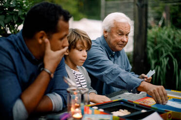 Multi-generation family playing board game while sitting at table in backyard - MASF16454