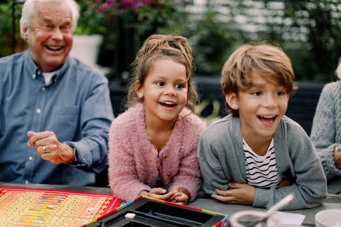 Lächelnde Enkelkinder spielen ein Brettspiel mit den Großeltern auf einem Tisch im Hinterhof - MASF16458