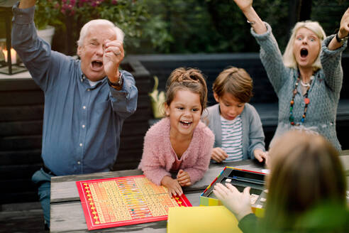 Lächelnde fröhliche Familie spielt Brettspiel, während sie am Tisch im Hinterhof sitzt - MASF16460