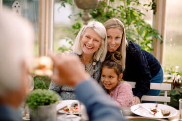 Älterer Mann fotografiert lächelnde Familie mit Mobiltelefon beim Mittagessen - MASF16471