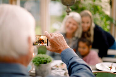 Senior man photographing happy family with mobile phone during lunch - MASF16474