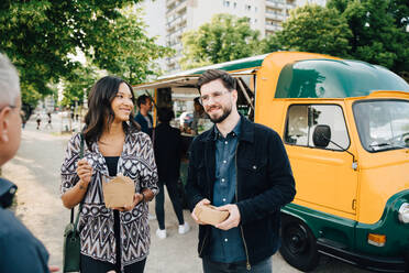 Smiling male and female friend with box standing against food truck - MASF16561