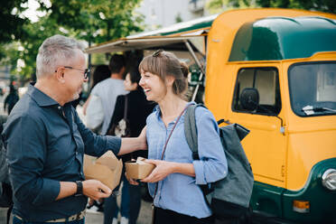 Happy male and female friends talking while standing with food box against commercial land vehicle - MASF16563