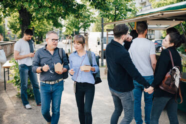 Male and female customer with box talking while couple holding hands and standing by food truck - MASF16564