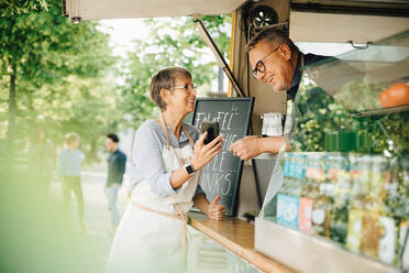 Female owner showing smart phone to male coworker while standing by food truck - MASF16573