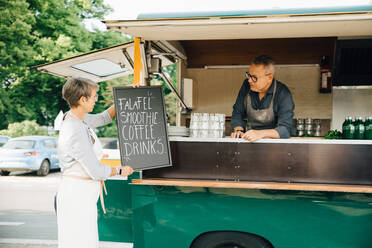 Female owner positioning board on concession stand while male coworker standing in food truck - MASF16575