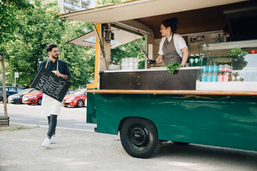 Male owner with blackboard looking at partner while walking by food truck - MASF16586