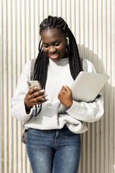 Junge Frau steht an einer Wand mit Handy und Laptop - VABF02591