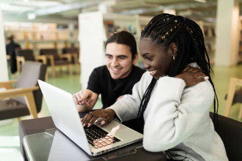 Zwei lächelnde Studenten benutzen einen Laptop in einer Bibliothek - VABF02604