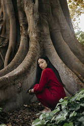 Schöne junge Frau in rotem Kleid, die an einem Baum mit großen Wurzeln hockt - TCEF00127