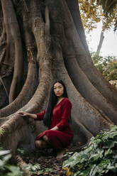 Schöne junge Frau in rotem Kleid, die an einem Baum mit großen Wurzeln hockt - TCEF00132