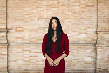 Portait of beautiful young woman wearing a red dress in front of a brick wall - TCEF00137