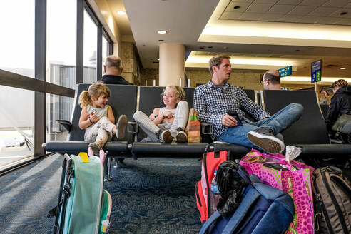 Zwei Mädchen und ihr Vater sitzen im Flughafenterminal und warten lachend - CAVF74286