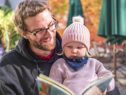 Porträt eines Vaters und eines Mädchens mit Kleinkind, das im Herbst in einem Café im Freien ein Buch liest - LAF02462