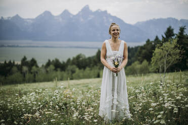 Die Braut steht in einem Blumenfeld mit Blumenstrauß und Bergen im Hintergrund - CAVF74653